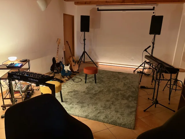 Kytary, syntezátor a piano kolem volného místa na koberci, s mikrofonem a reproduktory.